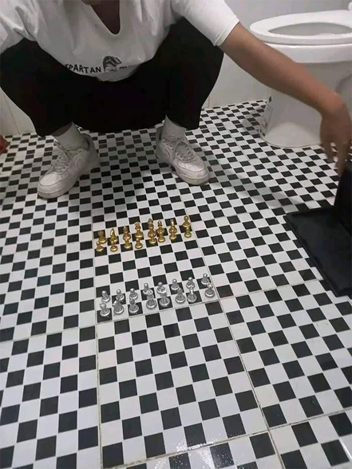 Person squatting in a bathroom with a chessboard floor and chess pieces set up, a cursed gaming setup example.
