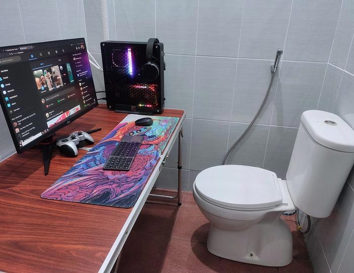 Minimal space small bathroom converted into a cursed gaming setup with PC, monitor, keyboard, and toilet side by side