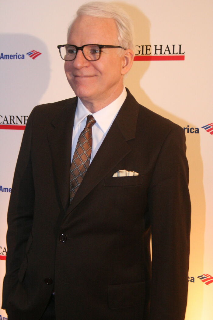Actor wearing glasses and a brown suit smiling at an event featuring some of the smartest celebrities known for intelligence and talent.