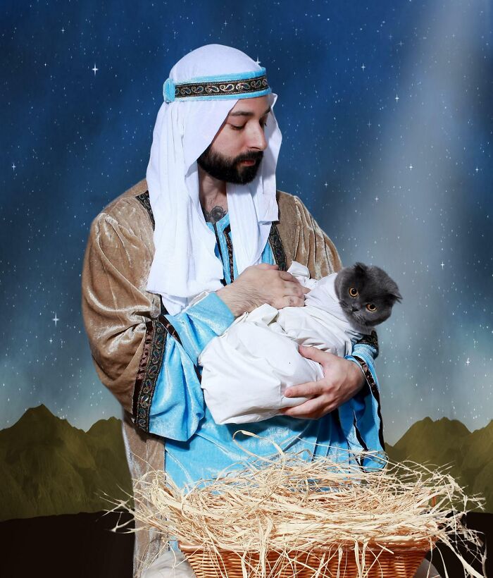 Man in a traditional outfit holding a grumpy cat wrapped in cloth, humorously styled as a nativity scene for a funny Christmas card.