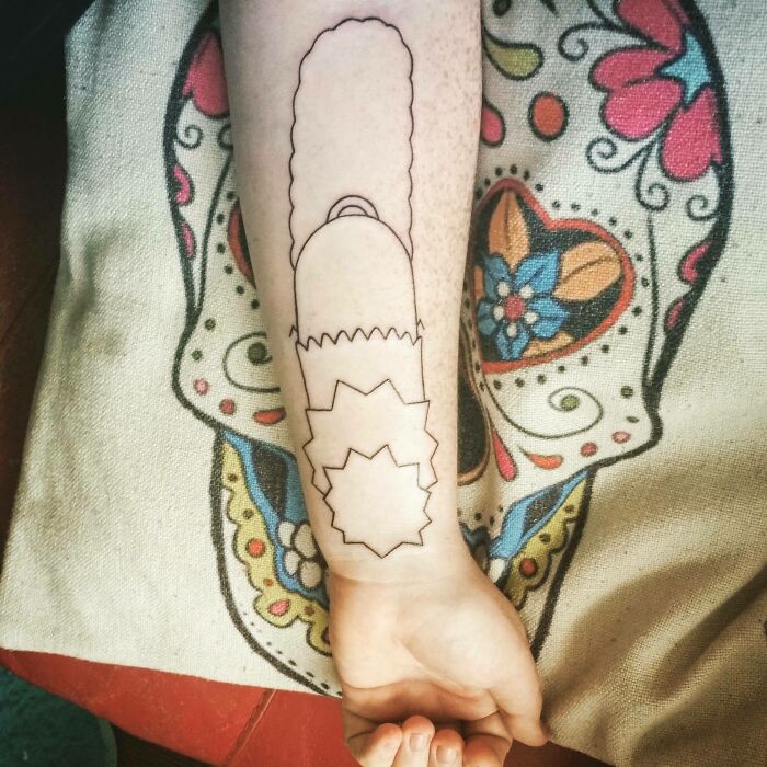 Cartoon tattoo outline of a chainsaw on an arm inspired by animated shows, with a colorful fabric in the background.