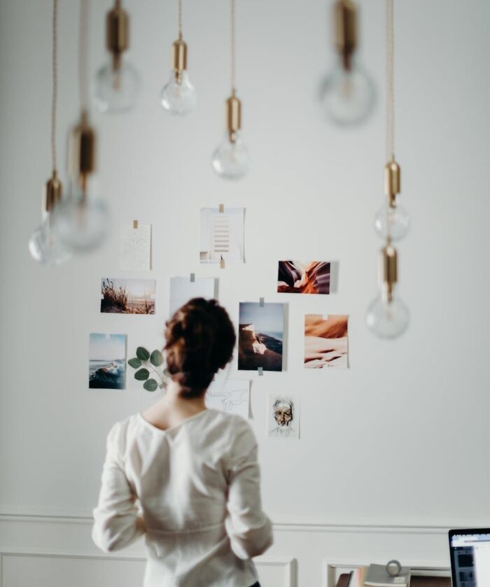 A woman planning cool New Year’s Eve activities while looking at photos on a wall in a cozy room with hanging light bulbs.