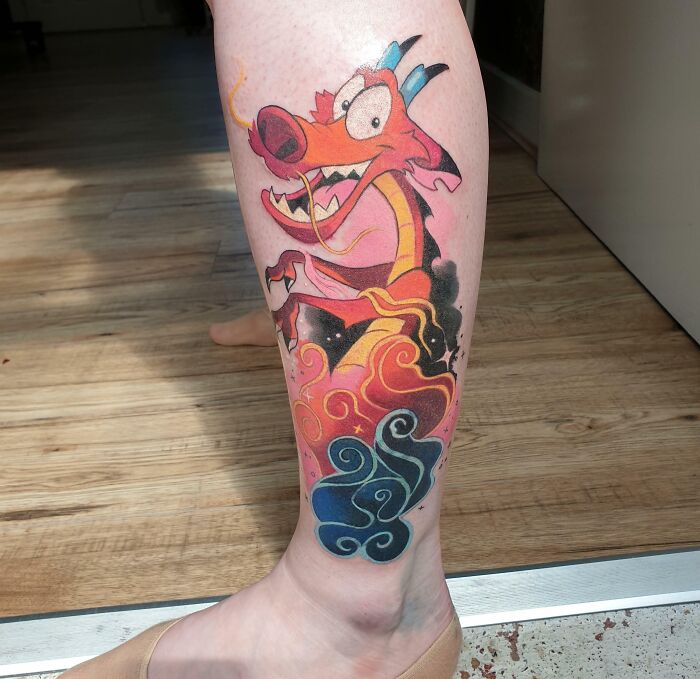 Colorful cartoon tattoo of a dragon character on a person's leg, featuring vibrant clouds.