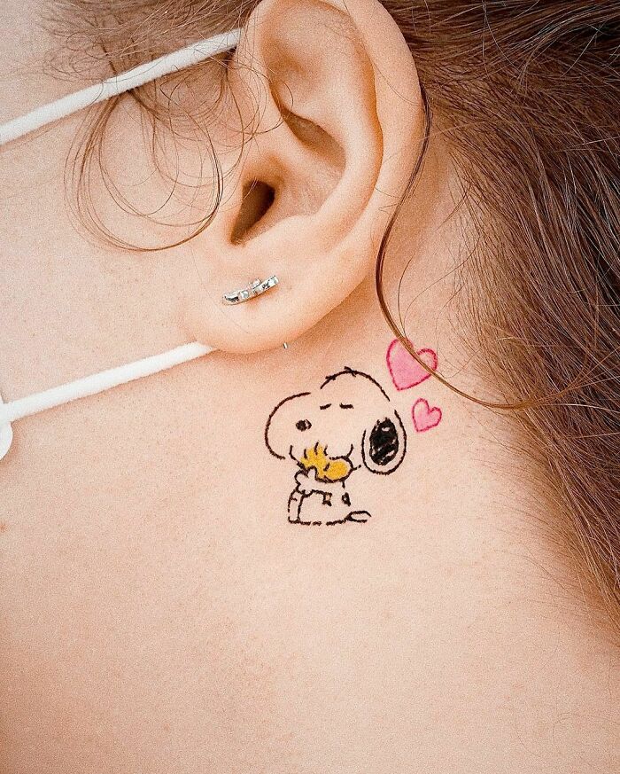 Cartoon tattoo of Snoopy and Woodstock hugging on ear, featuring pink hearts.