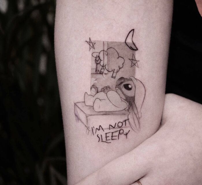 Cartoon tattoo of Stitch with insomnia, featuring stars and moon, on an arm.