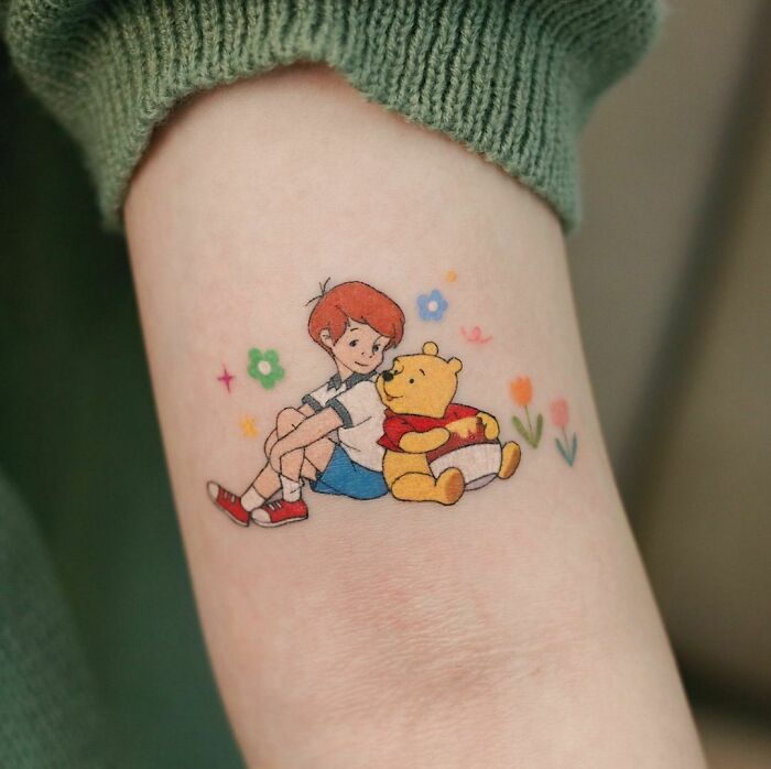 Cartoon tattoo featuring a boy Robin sitting with a teddy bear Pooh surrounded by colorful flowers on an arm.