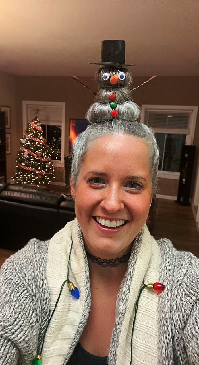 Person with a snowman hairdo and Christmas lights, smiling in front of a decorated tree, showcasing holiday humor.