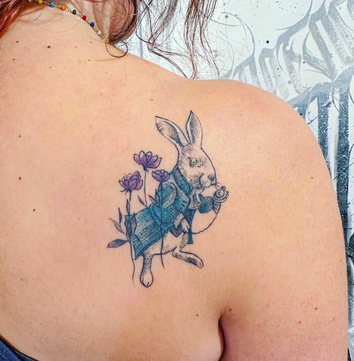 Cartoon tattoo of a rabbit in a blue coat holding a watch, surrounded by flowers, on someone's shoulder.