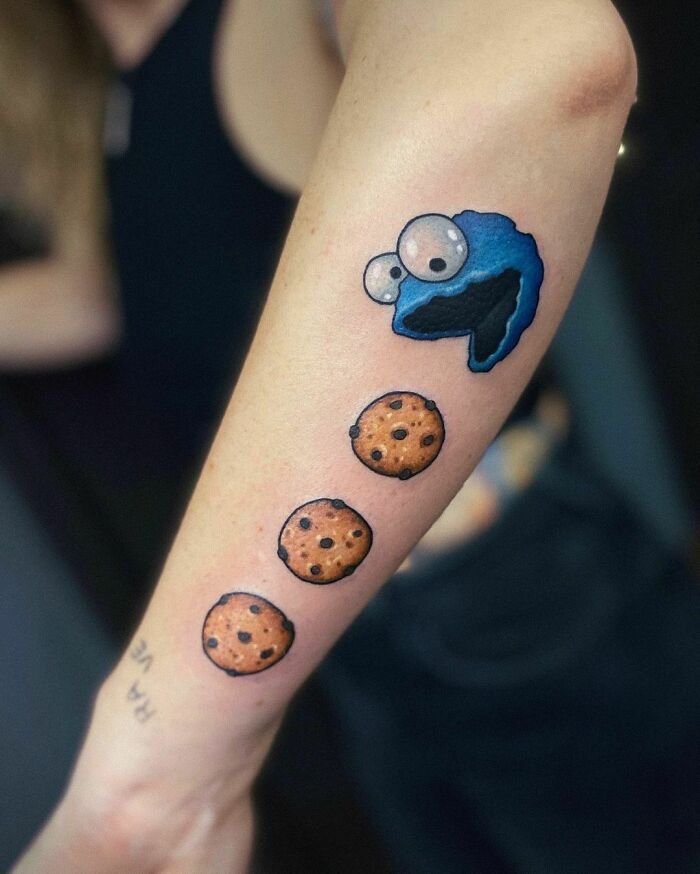 Cartoon tattoo of a blue character with cookies on forearm, inspired by childhood favorites.