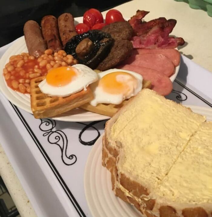 Full English breakfast with eggs, sausages, baked beans, mushrooms, bacon, and buttered toast on a white plate.
