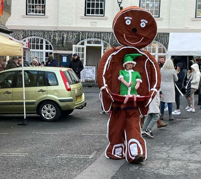Person in gingerbread man costume holding a small elf figure, adding humor to Christmas street scene.