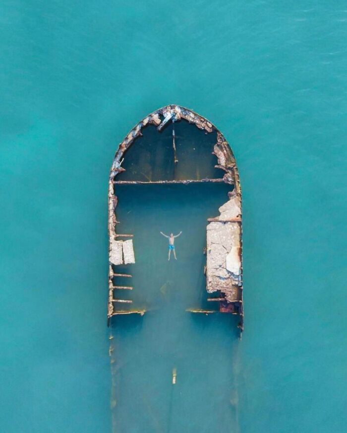Aerial view of a person floating inside a rusty, partially submerged shipwreck in calm ocean water, ocean frightening pics.