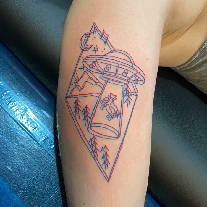 Trippy tattoo of a UFO abducting a cow, with mountains and trees in a surreal, colorful design on an arm.