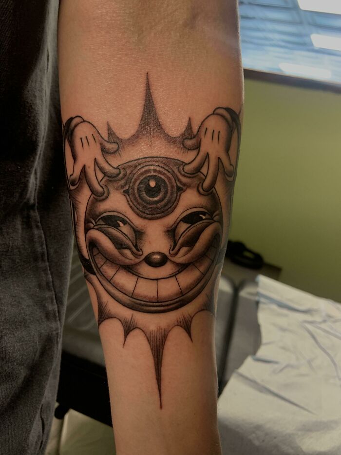 Trippy tattoo of a grinning sun with an eye and hands, creating an optical illusion on a forearm.