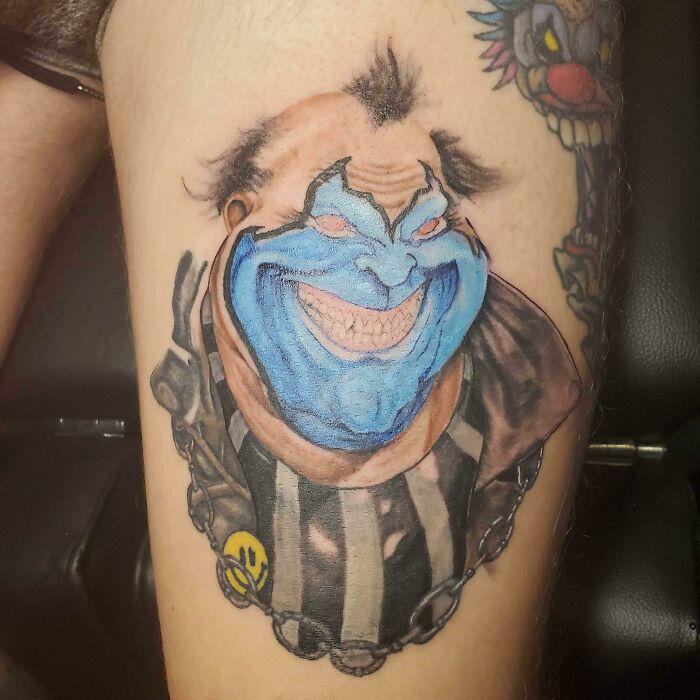 Trippy tattoo of an evil clown with a blue face inked on a leg.