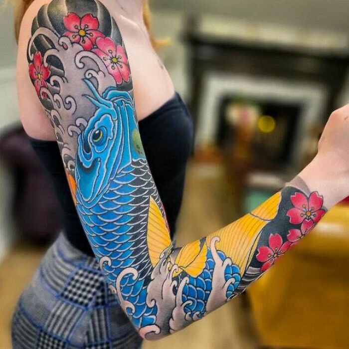 Colorful psychedelic tattoo sleeve featuring a blue koi fish and pink flowers on a woman's arm.