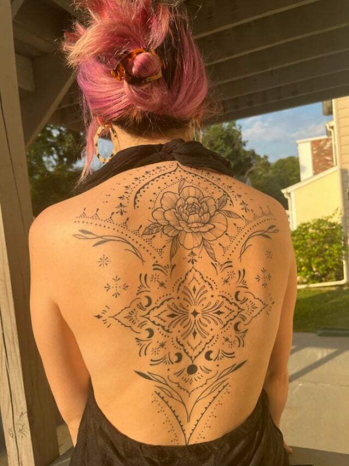 Intricate trippy tattoo design on a woman's back with floral and geometric patterns, under a pergola.