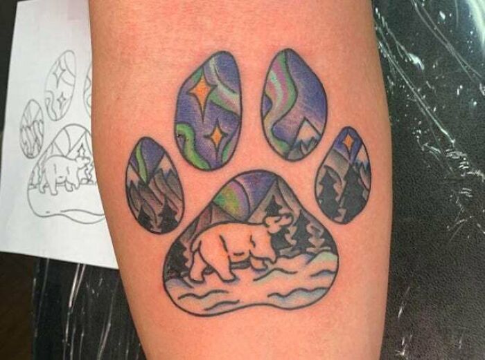 Trippy tattoo of a bear in a paw print design with a colorful aurora and mountain scenery.