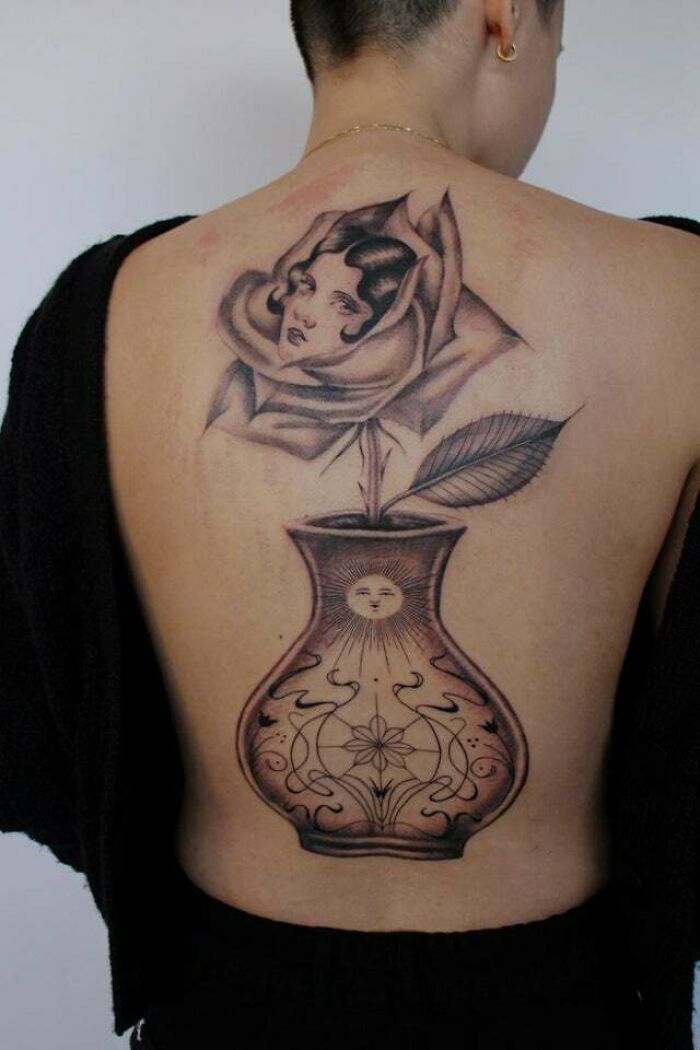 Trippy tattoo of a vase with a rose and a face on a person's back, featuring intricate designs.