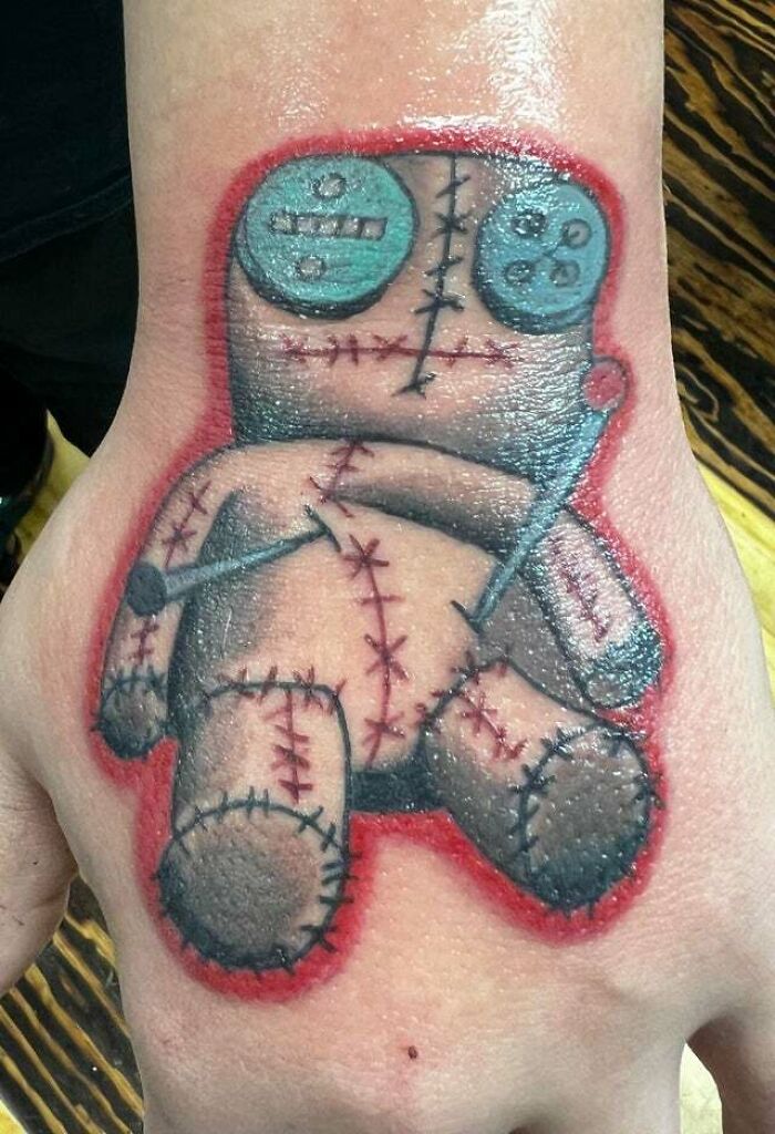 Trippy tattoo of a stitched doll with button eyes on a hand, featuring red and blue hues.