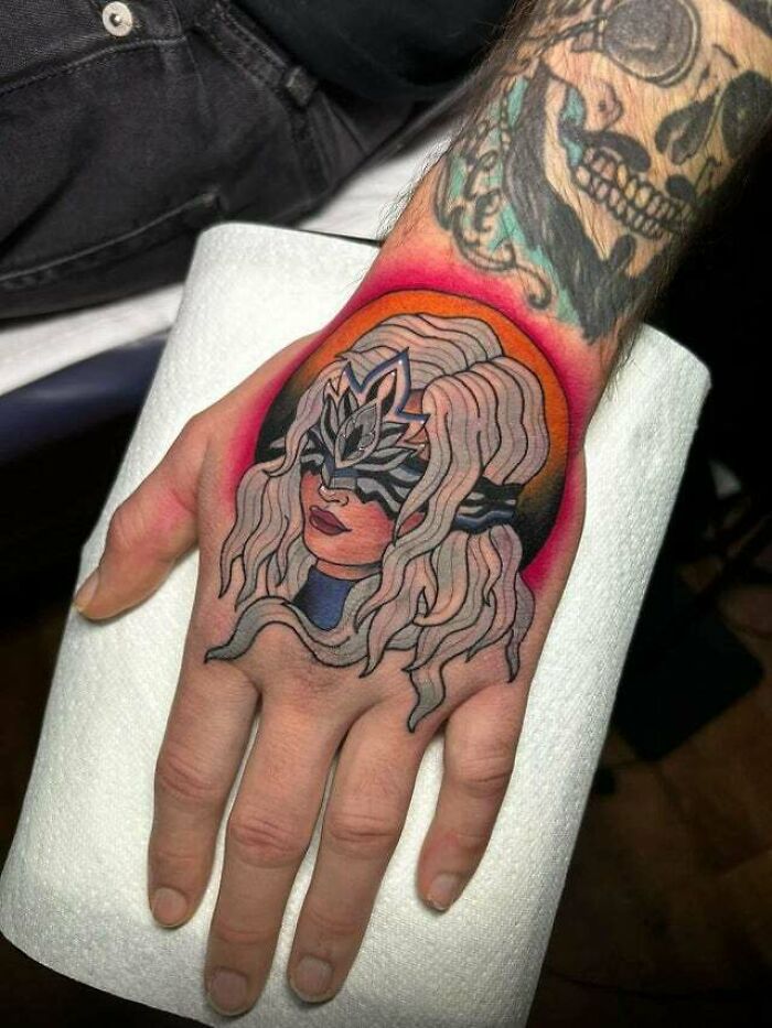 Trippy tattoo of a masked woman with flowing hair on a hand, featuring vibrant colors and abstract design.