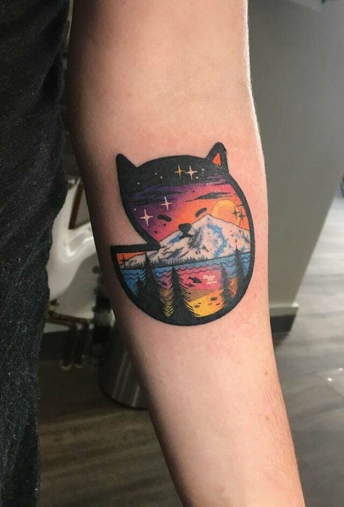 Trippy cat symbol tattoo with colorful nature scene on an arm.