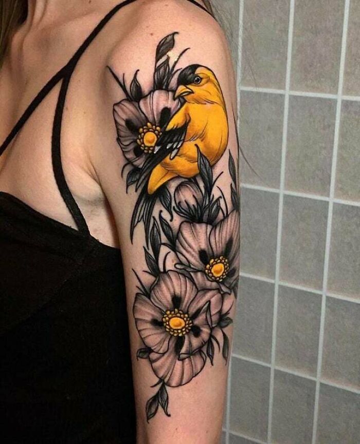 Trippy tattoo of a vibrant yellow bird with black wings perched on detailed flowers on an upper arm.