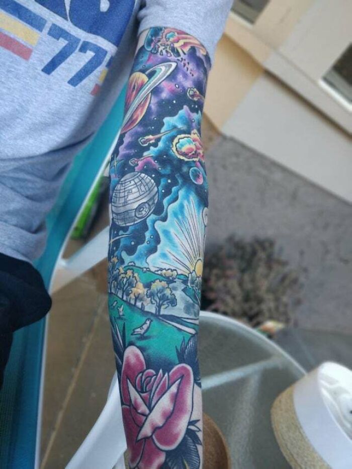 Colorful trippy tattoo of a cosmic scene with planets, stars, and a rose on an arm.