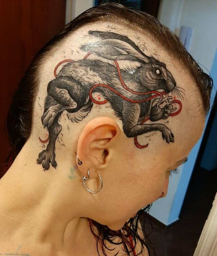 Side profile of a person with a trippy rabbit tattoo on their shaved scalp, featuring swirling red ribbons.