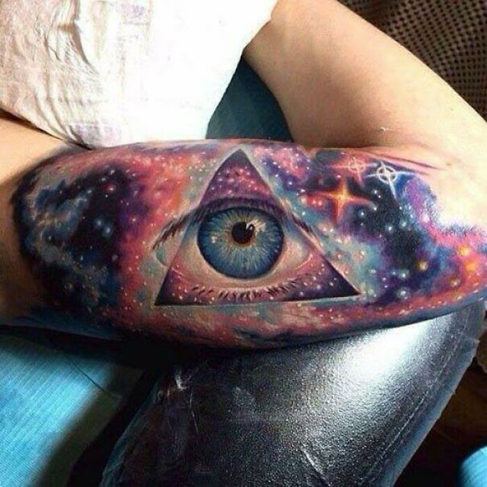 Colorful psychedelic art tattoo featuring an eye within a triangular galaxy design on an arm.