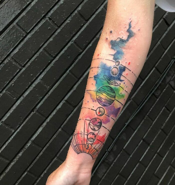 Trippy tattoo of planets and watercolor galaxy on a forearm with a brick background.