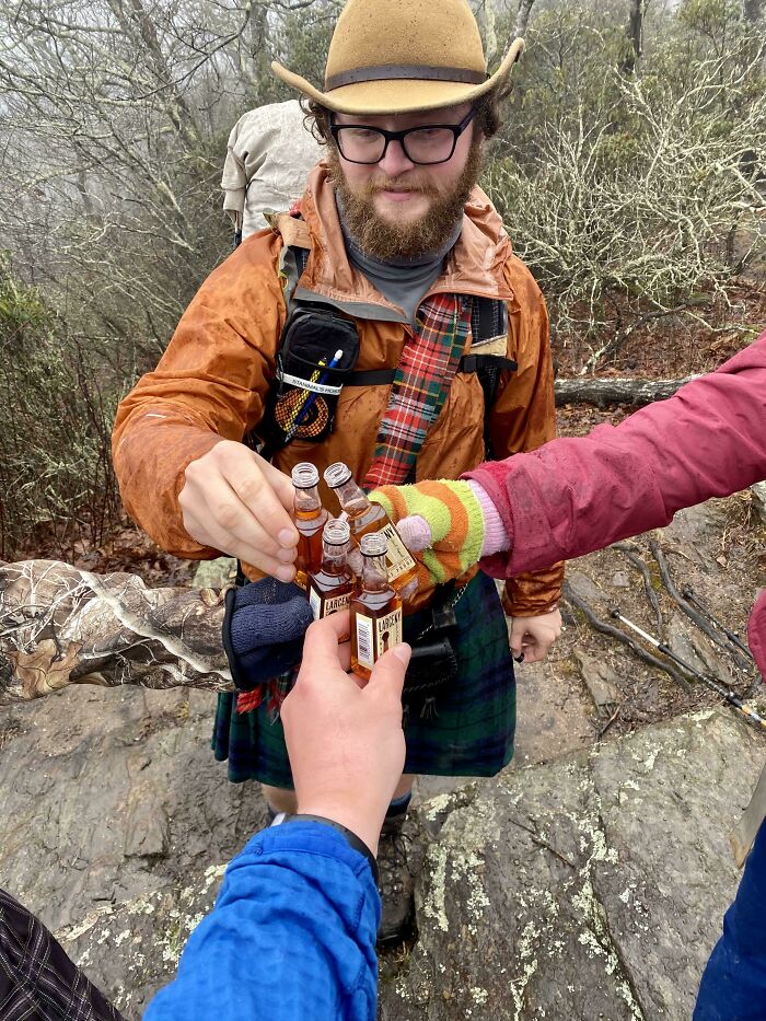 Group of hikers toasting with small bottles in a forest, showcasing perfectly camouflaged outdoor gear and clothing layers.