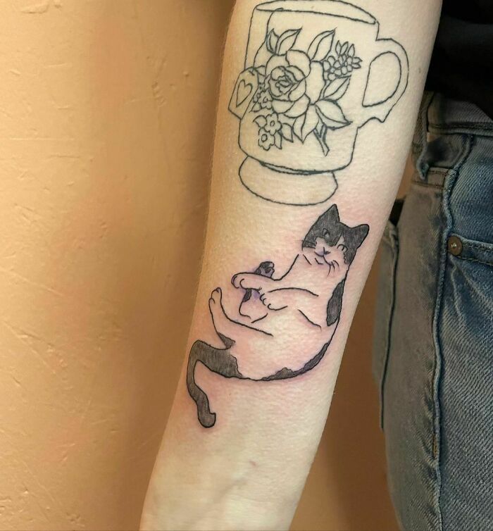 Tattoo of an adorable black and white cat on an arm, next to a floral mug design.