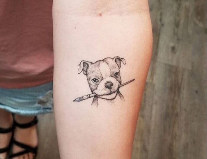 Tattoo of a dog holding a pencil, celebrating beautiful pet art on an arm.