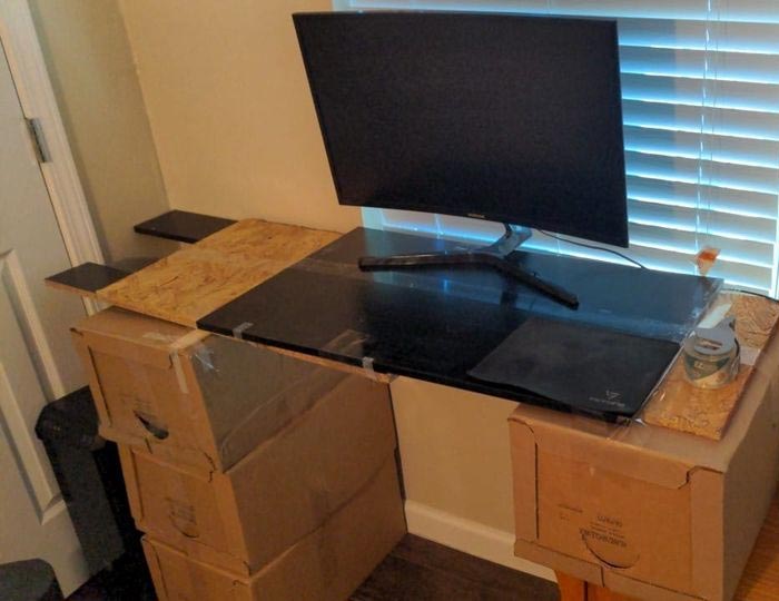 Gaming setup built with cardboard boxes and wooden boards holding a curved monitor in a small room.