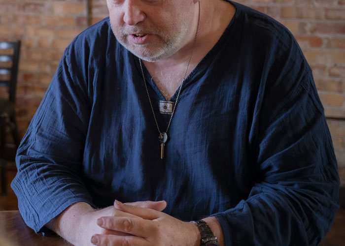Middle-aged man in dark shirt and key necklace sitting at a wooden table sharing disturbing things experienced at work.