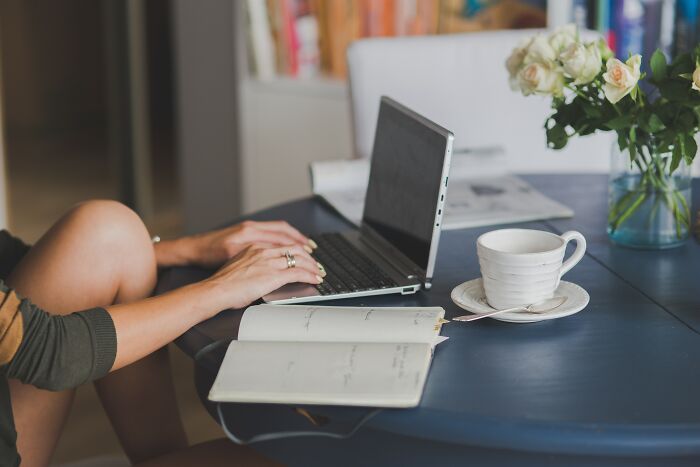 Person typing on a laptop at a blue table with an open notebook, cup of coffee, and vase of flowers nearby.