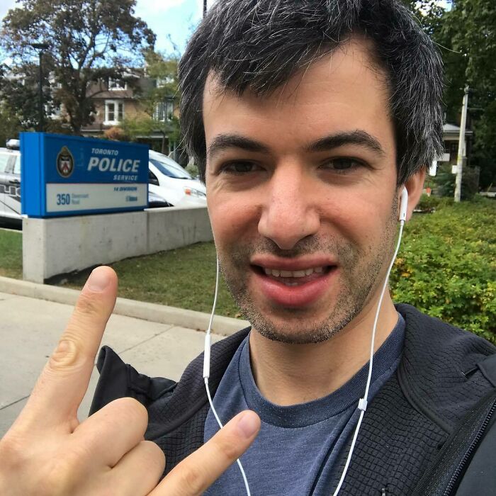 Man wearing earbuds, smiling and making a hand gesture outside Toronto Police, representing smartest celebrities.