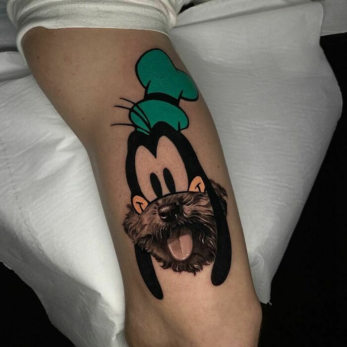 Cartoon tattoo of Goofy with a green hat on arm, blending with a realistic dog face.