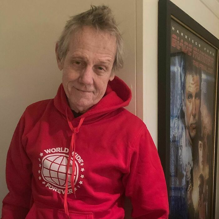 Older man wearing a red hoodie standing indoors next to framed movie poster, representing smartest celebrities and intelligence.
