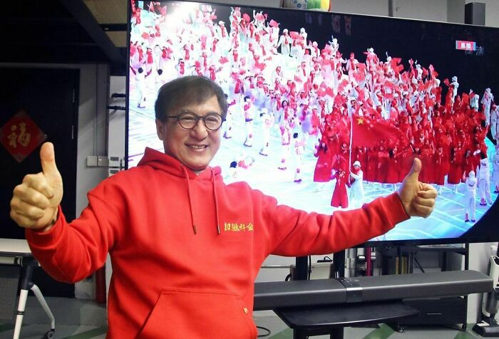 Celebrity in a red hoodie giving thumbs up, smiling in front of a large screen with a crowd, showcasing intelligent celebrities.