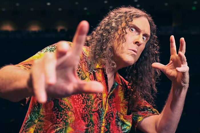 Comedian with long curly hair in colorful shirt striking a humorous pose, illustrating smartest celebrities intelligence.