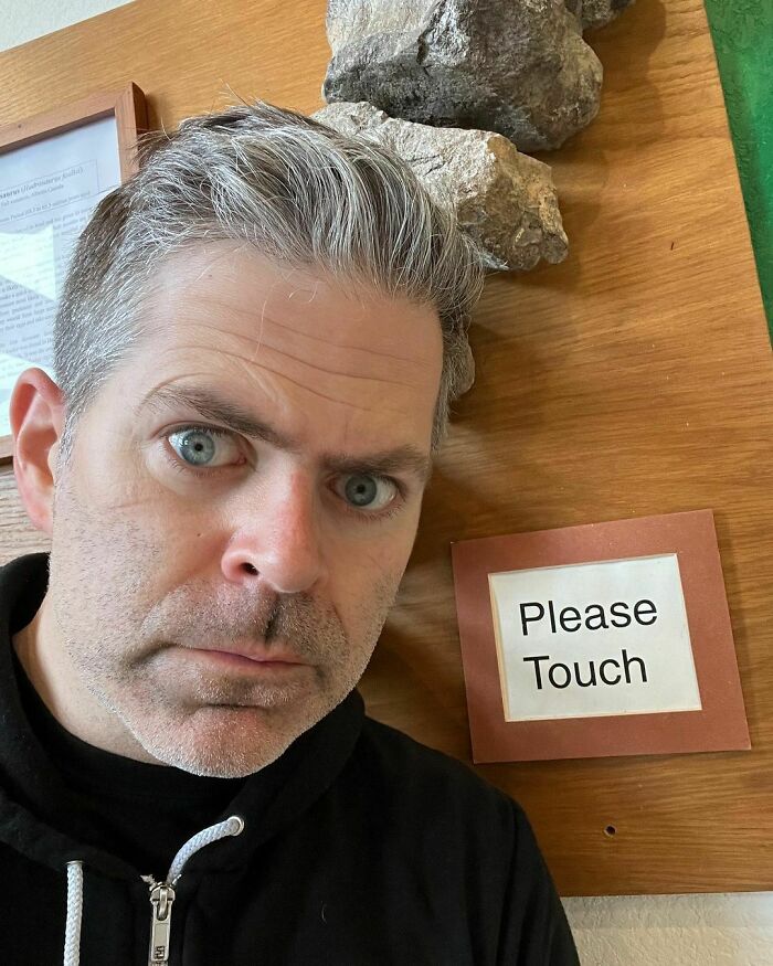 Man with gray hair and blue eyes wearing a black hoodie standing near a sign that says Please Touch, smart celebrities concept.