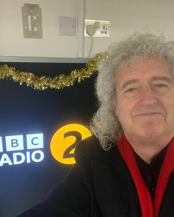 Older man with curly gray hair in a red scarf and black coat at a BBC Radio 2 studio, representing smartest celebrities.