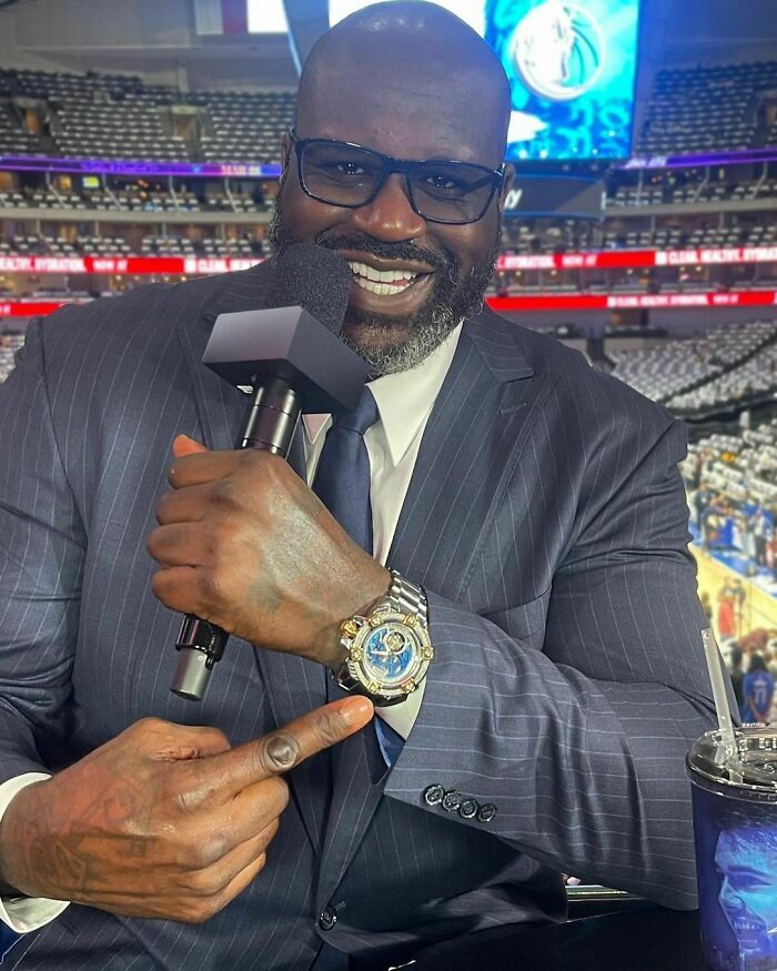 Celebrity in a pinstripe suit holding a microphone and pointing to his watch during a sporting event broadcast.