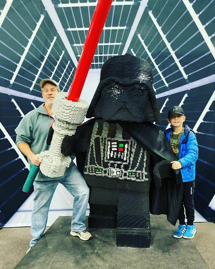 Life-sized Darth Vader LEGO build with red lightsaber, standing between an adult and a child at a LEGO art exhibit.