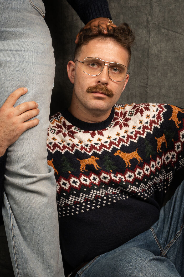 Man in 80's style glasses and sweater poses for a retro photoshoot, hand resting on nearby leg for an awkward effect.