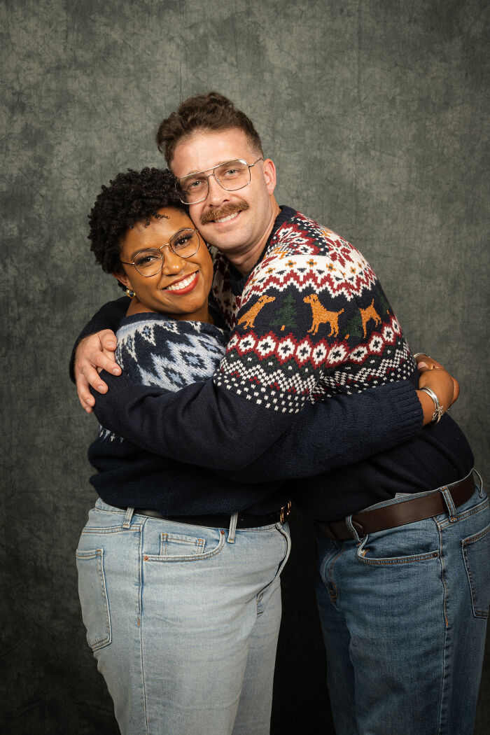 Couple embracing in an awkward 80's-themed photoshoot, wearing retro sweaters with a backdrop of a textured gray wall.