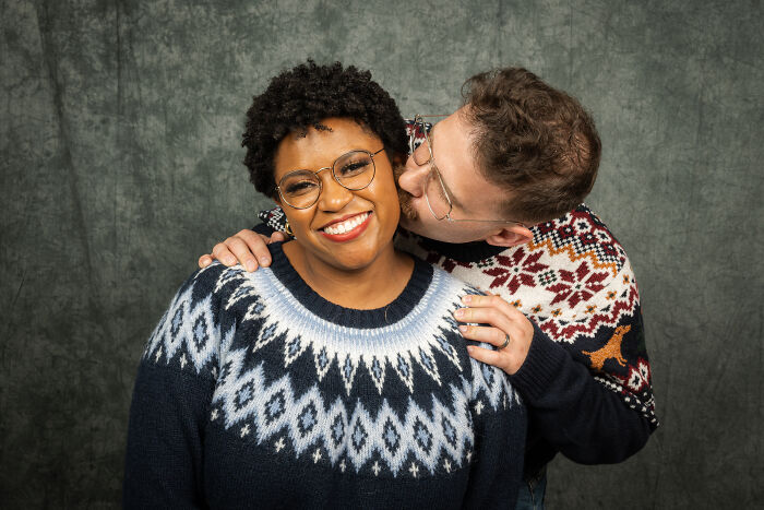 Couple in 80s-themed photoshoot, wearing retro sweaters; man kisses woman, both smiling joyfully for their anniversary.