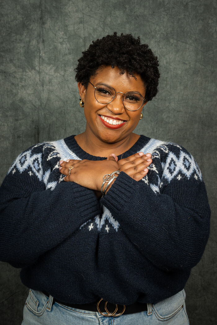 Person smiling in an 80's style photoshoot, wearing glasses and a patterned sweater, arms crossed over chest.